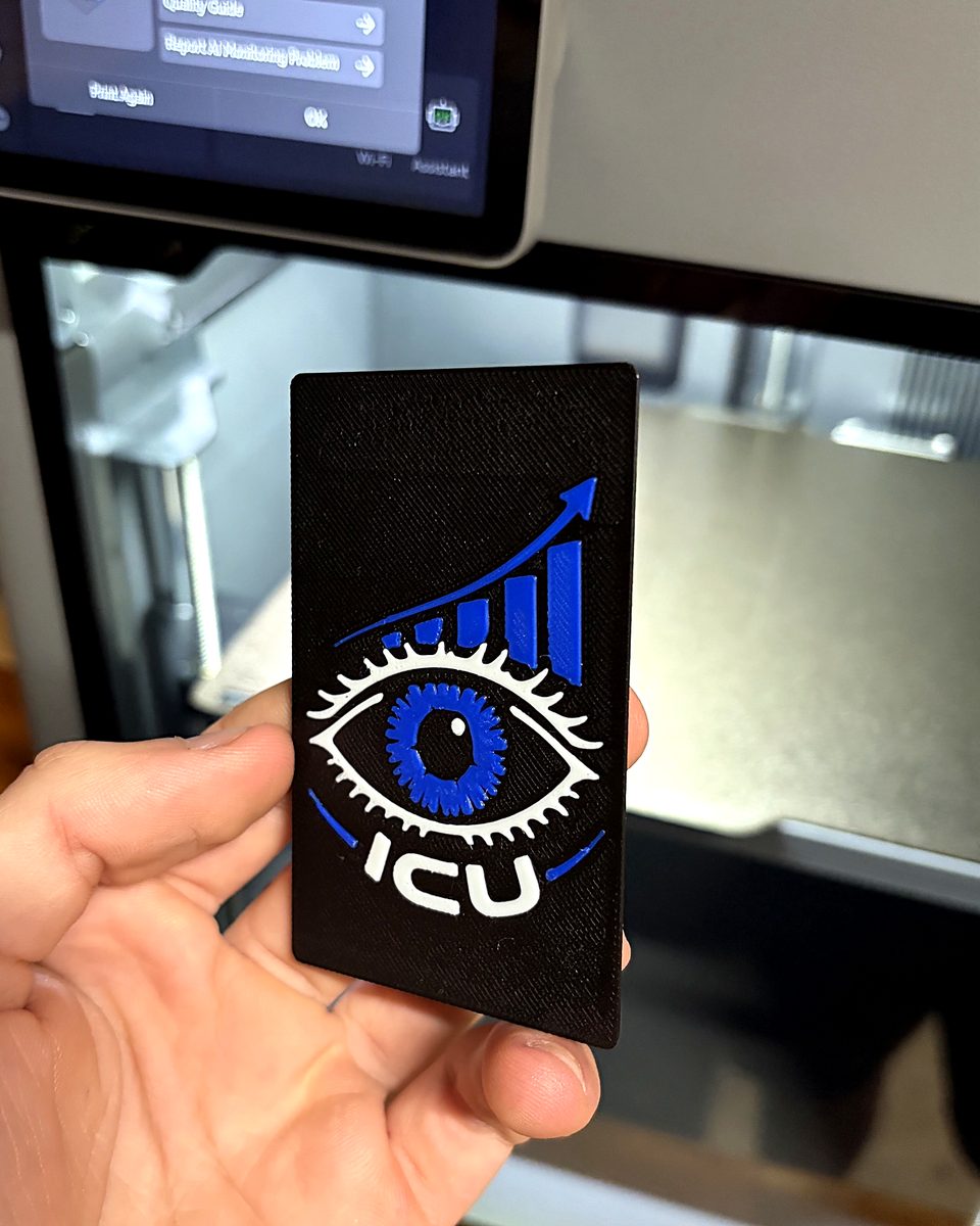 A black ICU card with a raised white eye and blue growth chart, held up in front of the 3D printer that just finished it